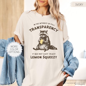 May include: Ivory-colored t-shirt with a raccoon graphic holding a lemon. The text reads "IN THE INTEREST OF FULL TRANSPARENCY" and "IT WAS NOT EASY PEASY LEMON SQUEEZY". The shirt has a relaxed fit and is a neutral color.
