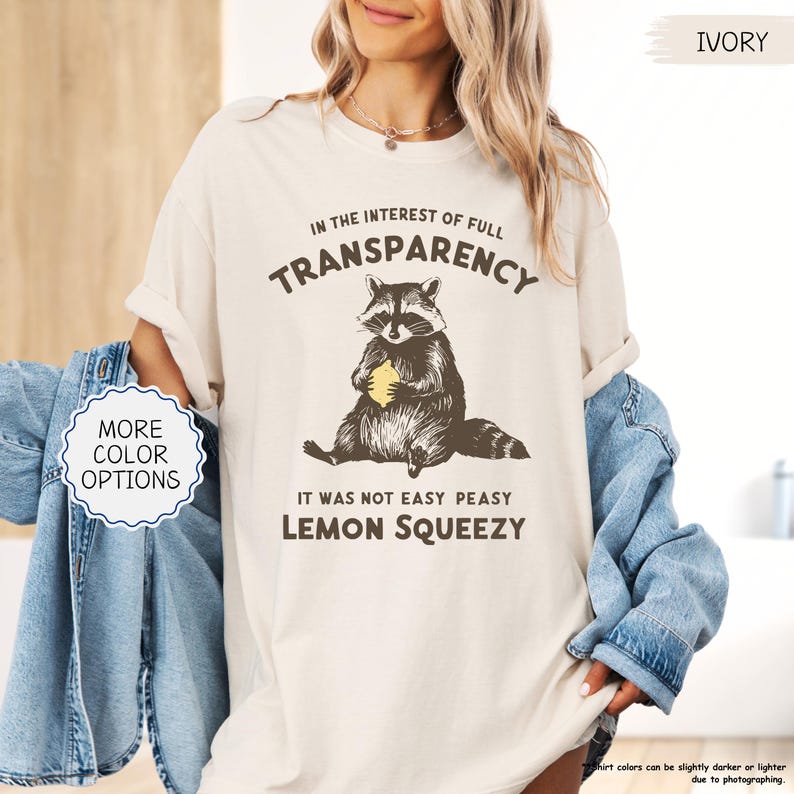 May include: Ivory-colored t-shirt with a raccoon graphic holding a lemon. The text reads "IN THE INTEREST OF FULL TRANSPARENCY" and "IT WAS NOT EASY PEASY LEMON SQUEEZY". The shirt has a relaxed fit and is a neutral color.