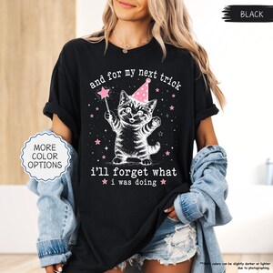 May include: A black t-shirt featuring a white graphic of a kitten wearing a pink party hat and holding a star-tipped wand. The text reads, and for my next trick i'll forget what i was doing, surrounded by small pink stars on the front of the shirt.