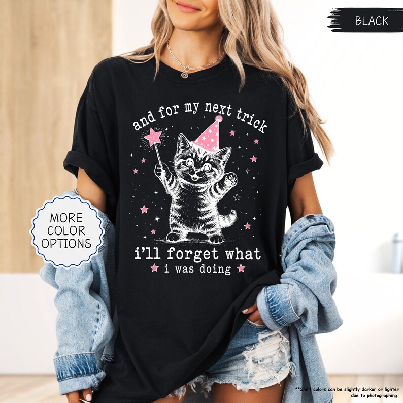 May include: A black t-shirt featuring a white graphic of a kitten wearing a pink party hat and holding a star-tipped wand. The text reads, and for my next trick i'll forget what i was doing, surrounded by small pink stars on the front of the shirt.