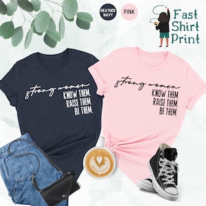 K&ouml;nnte beinhalten: Zwei T-Shirts mit dem Text "strong women know them. raise them. be them." bedruckt. Ein T-Shirt ist heidefarbenes Marineblau und das andere ist rosa.