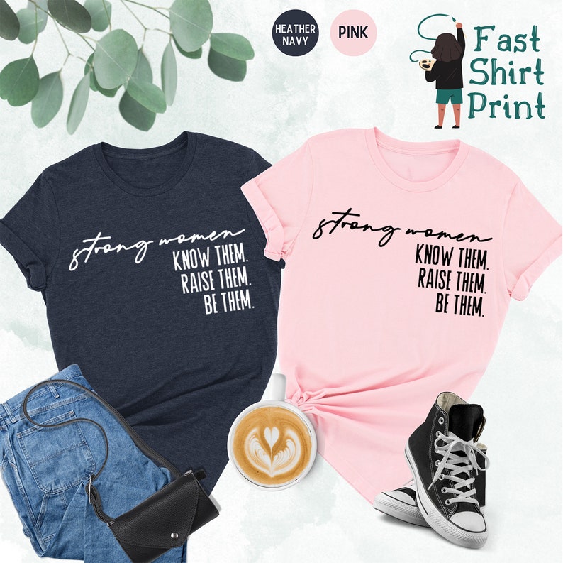 K&ouml;nnte beinhalten: Zwei T-Shirts mit dem Text "strong women know them. raise them. be them." bedruckt. Ein T-Shirt ist heidefarbenes Marineblau und das andere ist rosa.