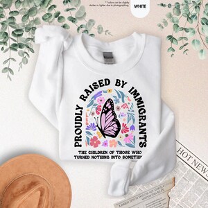 May include: White sweatshirt with the text "PROUDLY RAISED BY IMMIGRANTS" in black arched lettering. A butterfly with pink wings and a floral design is in the center. The text "THE CHILDREN OF THOSE WHO TURNED NOTHING INTO SOMETHI" is below.