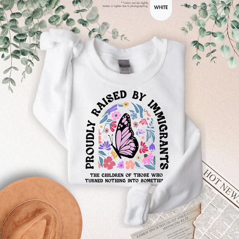 May include: White sweatshirt with the text "PROUDLY RAISED BY IMMIGRANTS" in black arched lettering. A butterfly with pink wings and a floral design is in the center. The text "THE CHILDREN OF THOSE WHO TURNED NOTHING INTO SOMETHI" is below.