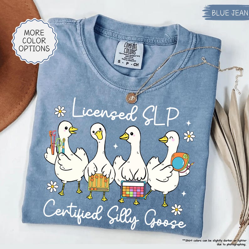 May include: A blue jean coloured t-shirt with the text "Licensed SLP Certified Silly Goose". The shirt features a graphic of four white cartoon geese holding various tools and instruments. A gold necklace is draped across the shirt.