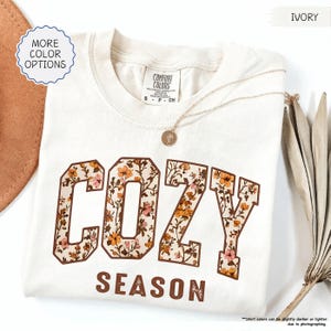 May include: Cream-coloured t-shirt featuring the words "COZY SEASON" in a floral design. The shirt has a crew neck and short sleeves. A gold necklace with a pendant is draped on the shirt. The words "MORE COLOR OPTIONS" are in a circle.