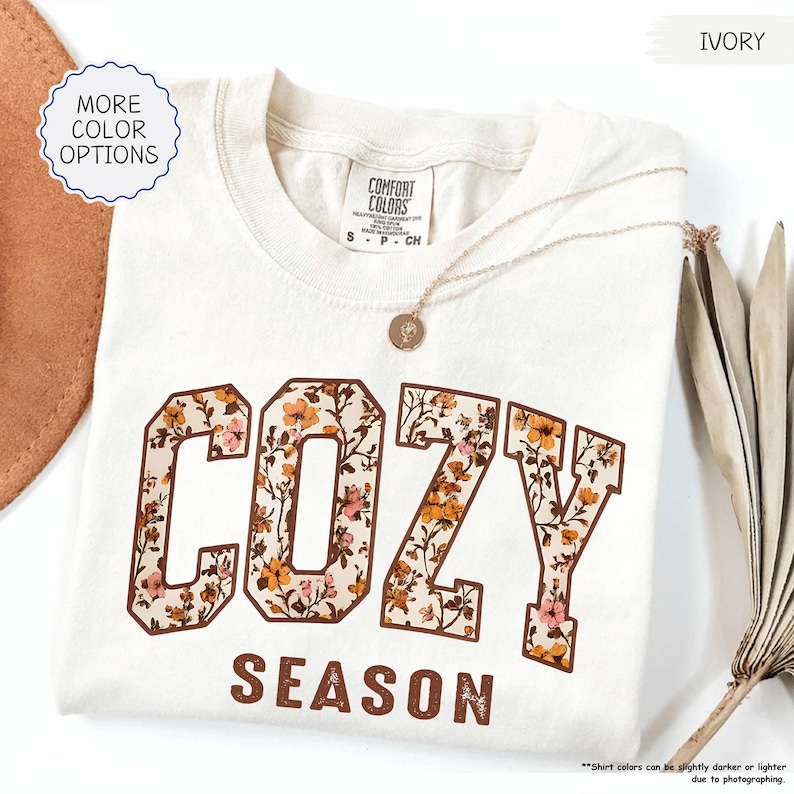 May include: Cream-coloured t-shirt featuring the words "COZY SEASON" in a floral design. The shirt has a crew neck and short sleeves. A gold necklace with a pendant is draped on the shirt. The words "MORE COLOR OPTIONS" are in a circle.