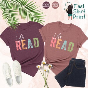 May include: Two heather maroon t-shirts with the text "Let's Read" in colorful, distressed letters. The shirts are laid out on a white background with white sneakers, a bouquet of white daisies, and a pair of blue jeans.