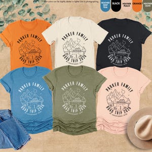May include: Several t-shirts in shades like orange, blue, and black, each displaying a mountain and lake design with the text "PARKER FAMILY ROAD TRIP 2026". The image also includes a pair of blue jeans and a hat. The shirts are in various sizes.