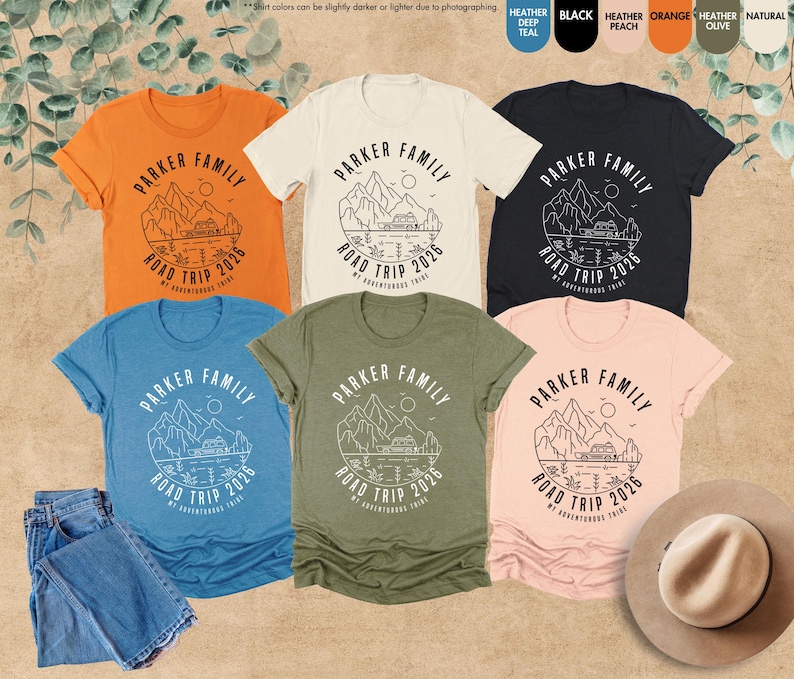 May include: Several t-shirts in shades like orange, blue, and black, each displaying a mountain and lake design with the text "PARKER FAMILY ROAD TRIP 2026". The image also includes a pair of blue jeans and a hat. The shirts are in various sizes.