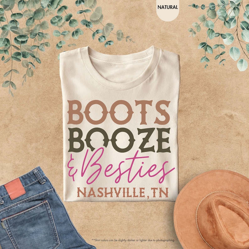 Pu&ograve; includere: Una maglietta color crema con le parole "BOOTS BOOZE & Besties NASHVILLE, TN" in lettere marroni, verdi e rosa. La maglietta &egrave; esposta con un paio di jeans blu e un cappello marrone.