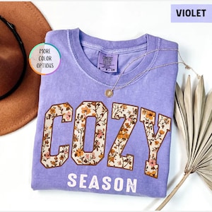 May include: A violet Comfort Colors t-shirt with the words "COZY SEASON" in a floral design. A gold necklace and a brown hat are next to the shirt. The shirt has a crew neck and short sleeves.