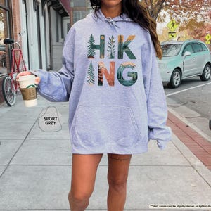 Pode incluir: Uma camisa de moletom cinza claro com capuz com a palavra "HIKING" impressa na frente em letras coloridas. As letras s&atilde;o cercadas por imagens de &aacute;rvores, montanhas e um p&ocirc;r do sol.