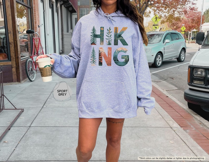 Pode incluir: Uma camisa de moletom cinza claro com capuz com a palavra "HIKING" impressa na frente em letras coloridas. As letras s&atilde;o cercadas por imagens de &aacute;rvores, montanhas e um p&ocirc;r do sol.