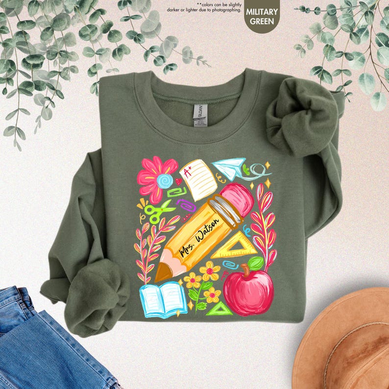 May include: Military green sweatshirt with a colorful school-themed graphic. The design includes a large pencil with "Mrs. Watson" printed on it, an apple, flowers, and school supplies. The sweatshirt is made of a soft material.