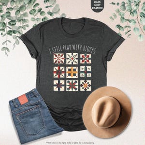 May include: A dark heather grey t-shirt with the text "I still play with blocks" printed on the front. The text is surrounded by a design of colorful quilt blocks.