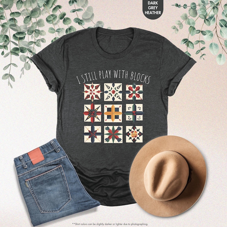 May include: A dark heather grey t-shirt with the text "I still play with blocks" printed on the front. The text is surrounded by a design of colorful quilt blocks.
