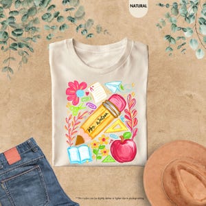 May include: Beige t-shirt with a colorful teacher-themed graphic. The design includes a large pencil with "Mrs. Watson" printed on it, a red apple, flowers, and school supplies. The shirt is displayed on a neutral surface.