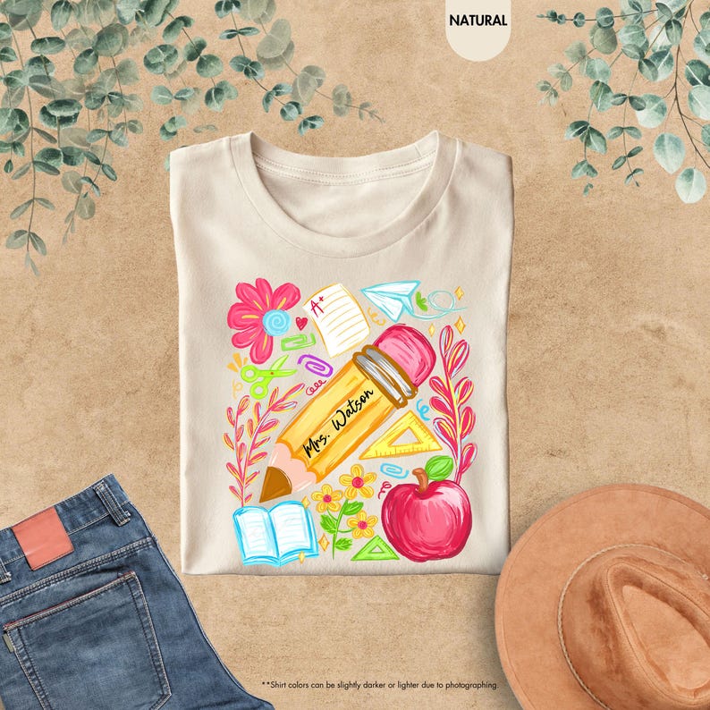 May include: Beige t-shirt with a colorful teacher-themed graphic. The design includes a large pencil with "Mrs. Watson" printed on it, a red apple, flowers, and school supplies. The shirt is displayed on a neutral surface.