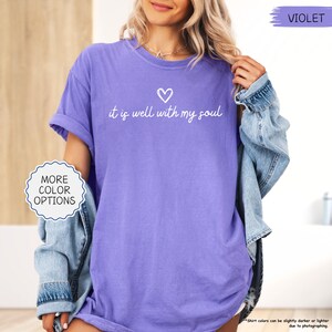 K&ouml;nnte beinhalten: Ein violettes T-Shirt mit dem Schriftzug "it is well with my soul" und einem Herz. Das Shirt hat hochgekrempelte &Auml;rmel und eine lockere Passform. Das Bild zeigt auch eine Jeansjacke und den Text "More Color Options".