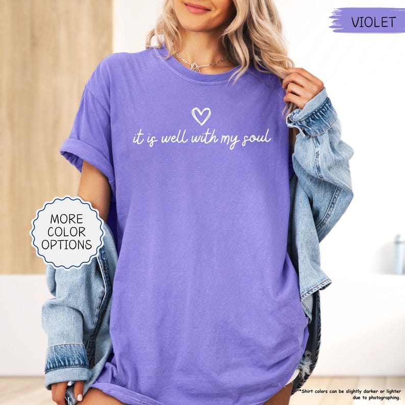 K&ouml;nnte beinhalten: Ein violettes T-Shirt mit dem Schriftzug "it is well with my soul" und einem Herz. Das Shirt hat hochgekrempelte &Auml;rmel und eine lockere Passform. Das Bild zeigt auch eine Jeansjacke und den Text "More Color Options".