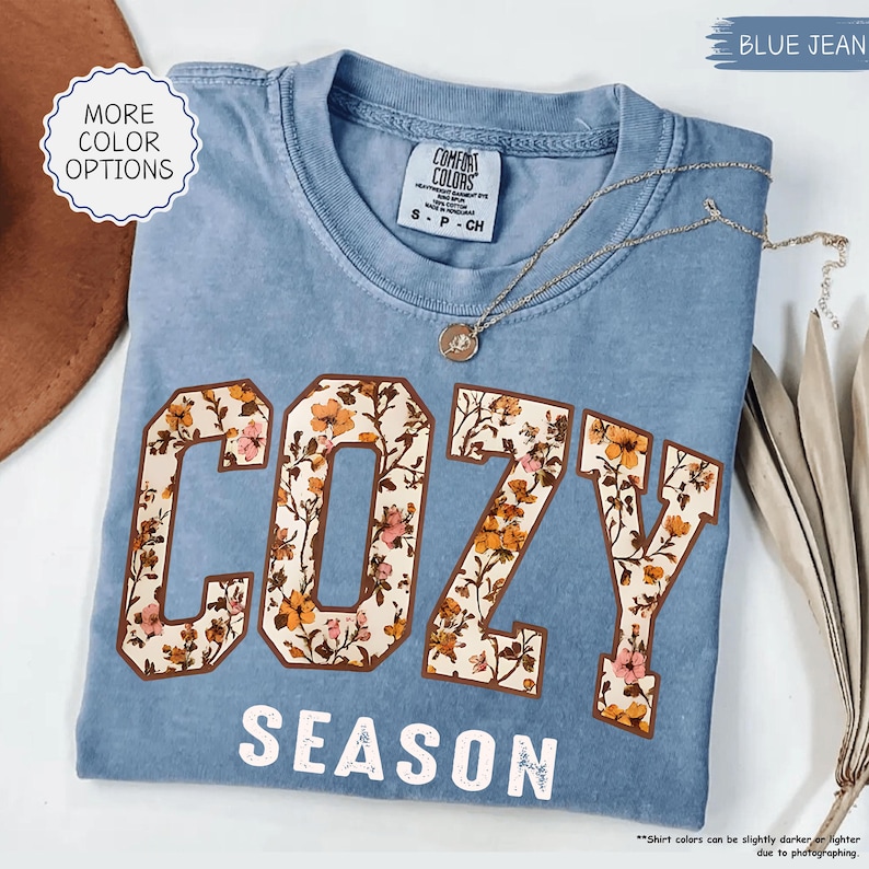 May include: A blue jean-coloured t-shirt with the words "COZY SEASON" in white. The letters of "COZY" are filled with a floral pattern in shades of orange, pink, and brown. A silver necklace with a pendant rests on the shirt.