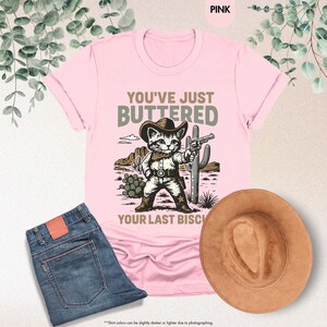 May include: Pale pink t-shirt featuring a cartoon cat in a cowboy outfit, holding a gun, with the text "YOU'VE JUST BUTTERED YOUR LAST BISCUIT". A pair of blue jeans and a brown cowboy hat are also visible.