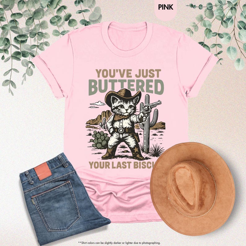 May include: Pale pink t-shirt featuring a cartoon cat in a cowboy outfit, holding a gun, with the text "YOU'VE JUST BUTTERED YOUR LAST BISCUIT". A pair of blue jeans and a brown cowboy hat are also visible.