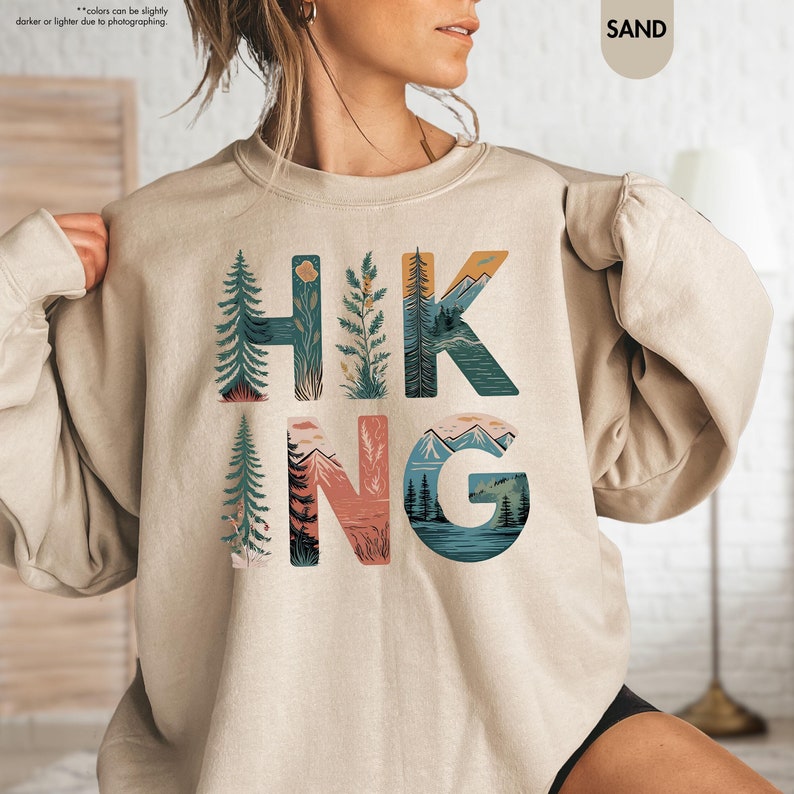 Pode incluir: Uma camisa de moletom bege com a palavra "HIKING" impressa na frente. As letras s&atilde;o compostas por ilustra&ccedil;&otilde;es de montanhas, &aacute;rvores e outras cenas da natureza.