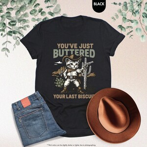 May include: Black t-shirt featuring a cat in a cowboy hat holding a gun, with the text "YOU'VE JUST BUTTERED YOUR LAST BISCUIT." The image also shows blue jeans and a brown cowboy hat.