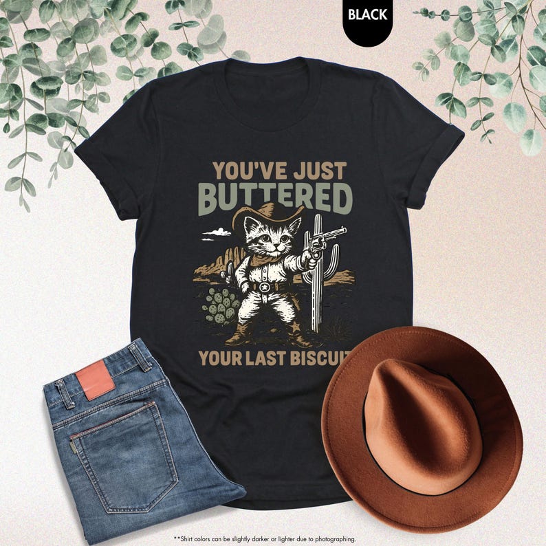 May include: Black t-shirt featuring a cat in a cowboy hat holding a gun, with the text "YOU'VE JUST BUTTERED YOUR LAST BISCUIT." The image also shows blue jeans and a brown cowboy hat.