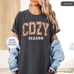 May include: Dark grey t-shirt featuring the words "COZY SEASON". "COZY" is in large, floral-patterned block letters, and "SEASON" is in white, sans-serif font. The shirt has rolled-up sleeves. The shirt is a Pepper color.