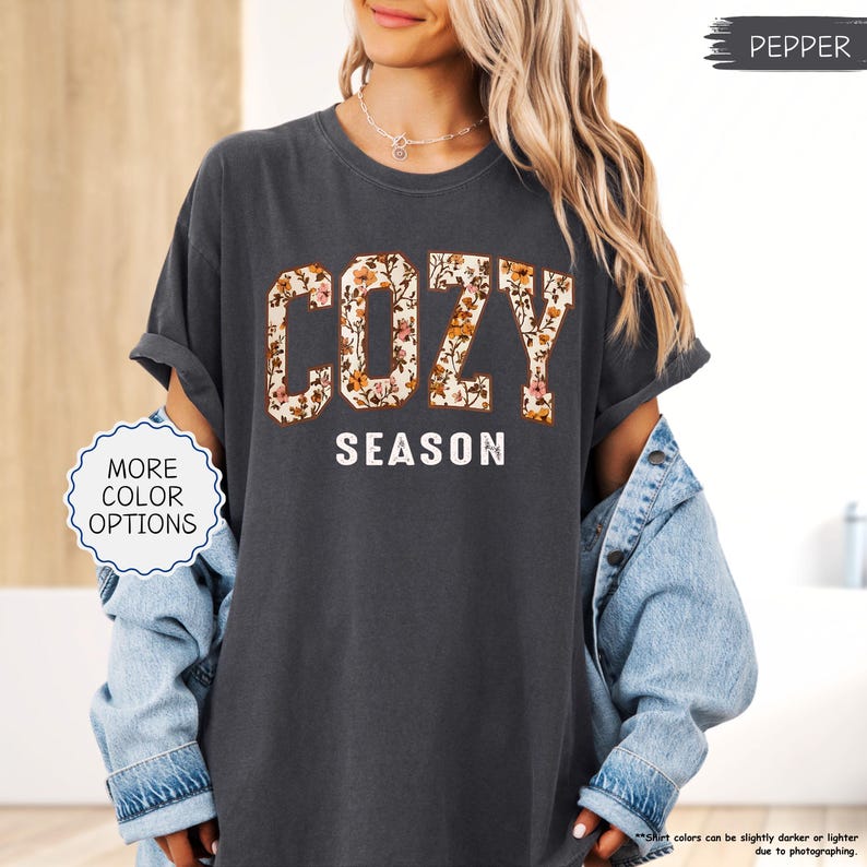 May include: Dark grey t-shirt featuring the words "COZY SEASON". "COZY" is in large, floral-patterned block letters, and "SEASON" is in white, sans-serif font. The shirt has rolled-up sleeves. The shirt is a Pepper color.