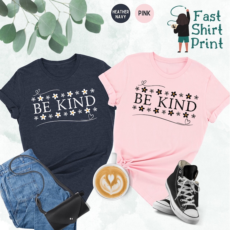 K&ouml;nnte beinhalten: Zwei T-Shirts mit dem Text "BE KIND" umgeben von kleinen Blumengrafiken. Die Shirts sind in Heather Navy und Pink.