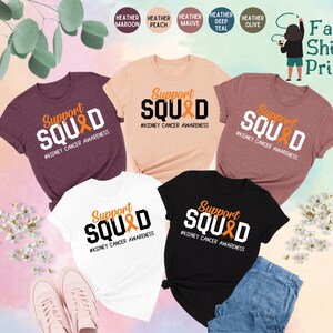May include: Five t-shirts in different colors with the text "Support SQUAD #KIDNEY CANCER AWARENESS" printed on them. The shirts are displayed on a white background with flowers and a pair of pink shoes.