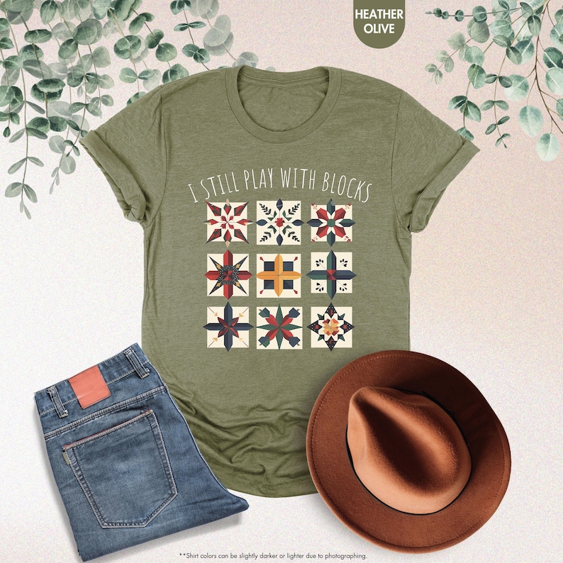 May include: A green t-shirt with the text "I Still Play With Blocks" and a design of colorful quilt blocks. The shirt is on a white background with a pair of blue jeans and a brown hat.