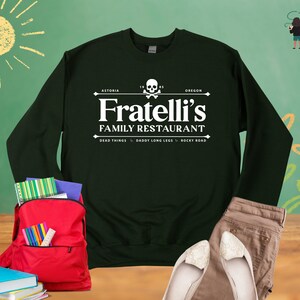 May include: A dark green sweatshirt with a white graphic print of a restaurant logo. The logo says "Fratelli's Family Restaurant" with the words "Astoria" and "Oregon" above and "Dead Things, Daddy Long Legs, Rocky Road" below.