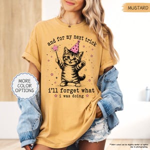 May include: A mustard yellow short-sleeve t-shirt featuring a graphic of a tabby kitten wearing a pink party hat and holding a magic wand. The text reads, "and for my next trick i'll forget what i was doing," surrounded by small pink stars.