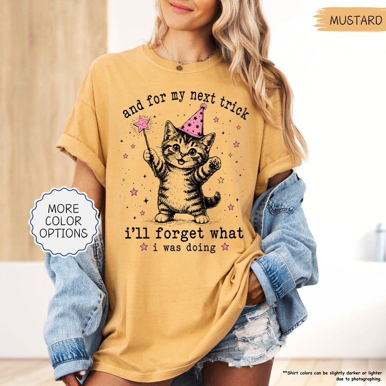 May include: A mustard yellow short-sleeve t-shirt featuring a graphic of a tabby kitten wearing a pink party hat and holding a magic wand. The text reads, "and for my next trick i'll forget what i was doing," surrounded by small pink stars.