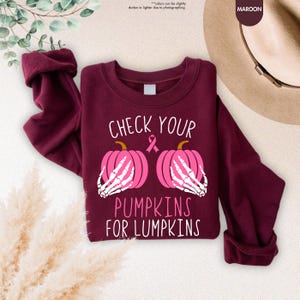 以下が含まれることがあります： 「CHECK YOUR PUMPKINS FOR LUMPKINS」の文字が入った、マルーン色のスウェットシャツ。デザインは、2つのピンク色のカボチャと、骸骨の手、ピンクのリボンが特徴です。スウェットシャツは無地のマルーン色です。