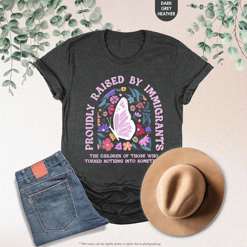 May include: Dark grey heather t-shirt with the text "PROUDLY RAISED BY IMMIGRANTS" in pink, featuring a butterfly and floral design. The shirt is paired with blue jeans and a tan hat.
