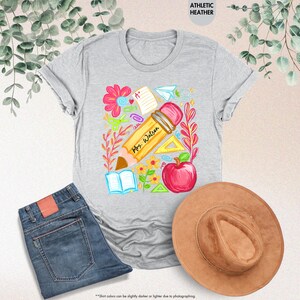 May include: Athletic heather gray t-shirt with a colorful teacher-themed graphic. The design includes a large pencil with "Mrs. Watson" written on it, an apple, flowers, and school supplies. A pair of blue jeans and a brown hat are also in the image.
