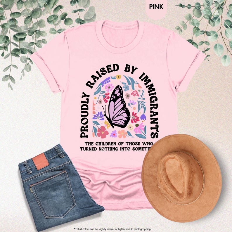 May include: Pink t-shirt with the text "PROUDLY RAISED BY IMMIGRANTS" and a butterfly graphic. The shirt is paired with blue jeans and a brown hat. The text "THE CHILDREN OF THOSE WHO TURNED NOTHING INTO SOMETHING" is also on the shirt.