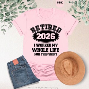 Puede incluir: Camiseta rosa claro con el texto negro "RETIRED 2026 I WORKED MY WHOLE LIFE FOR THIS SHIRT". La camiseta se combina con vaqueros azules y un sombrero marr&oacute;n. La palabra "PINK" est&aacute; en la esquina superior derecha.