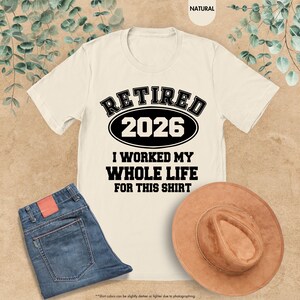 Puede incluir: Camiseta color crema con el texto "RETIRED 2026" en negrita negra. Debajo, se lee "I WORKED MY WHOLE LIFE FOR THIS SHIRT". La imagen tambi&eacute;n incluye unos vaqueros azules y un sombrero marr&oacute;n.