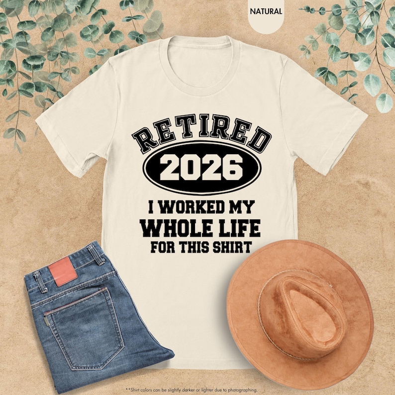 Puede incluir: Camiseta color crema con el texto "RETIRED 2026" en negrita negra. Debajo, se lee "I WORKED MY WHOLE LIFE FOR THIS SHIRT". La imagen tambi&eacute;n incluye unos vaqueros azules y un sombrero marr&oacute;n.