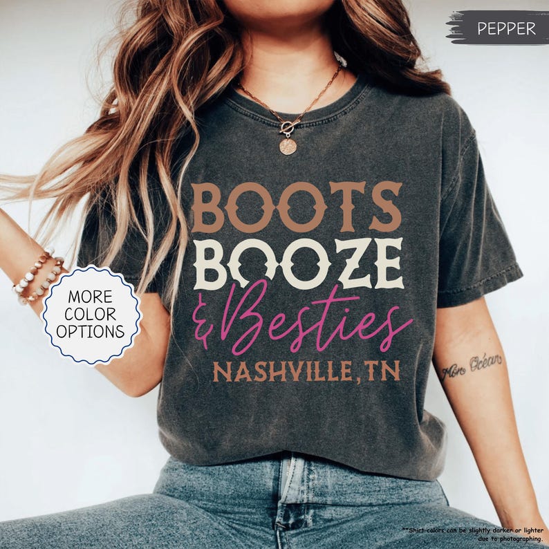 Pu&ograve; includere: T-shirt grigio scuro con la scritta "BOOTS BOOZE & Besties" in marrone, bianco e rosa. Il testo "NASHVILLE, TN" &egrave; sotto. La maglietta ha un aspetto vintage e usurato.