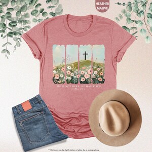 May include: A heather mauve t-shirt with a graphic of a cross on a hill with flowers. The text "HE IS NOT HERE. HE HAS RISEN. Matthew 28:6" is printed below the graphic.