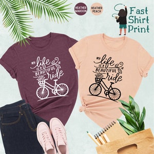 May include: Two t-shirts with the text "My life is a beautiful ride" and a bicycle graphic. The shirt on the left is heather maroon and the shirt on the right is heather peach.