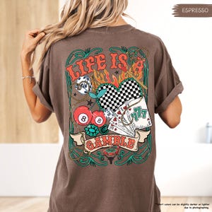 Puede incluir: Una camiseta marr&oacute;n con un estampado gr&aacute;fico de un coraz&oacute;n con un patr&oacute;n a cuadros, cerezas, dados, cartas de juego y el texto "Life is a Gamble".